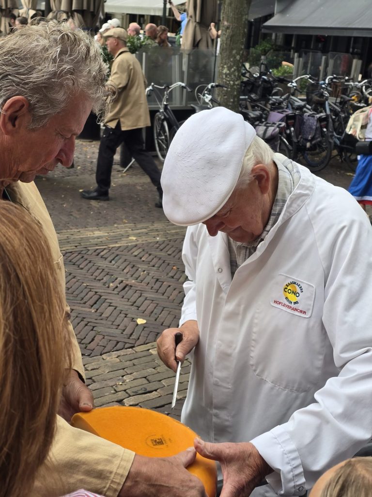 Cheese inspector in a white coat sampling a cheese wheel in front of guests at the Alkmaar market