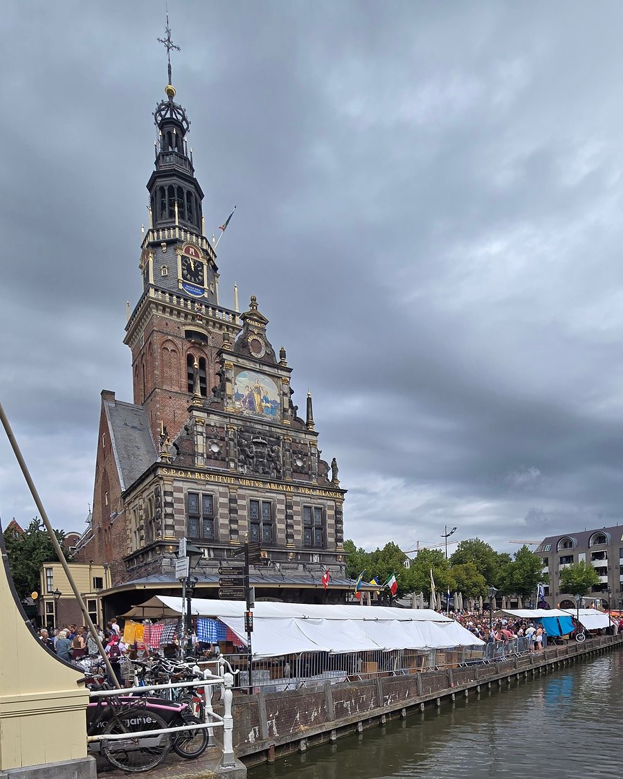 The Waag in Alkmaar, a brick and stone weigh house with a tall clock tower, seen across a canal from a drawbridge, with white market tents, parked bicycles and a crowd along the quay under a grey cloudy sky.