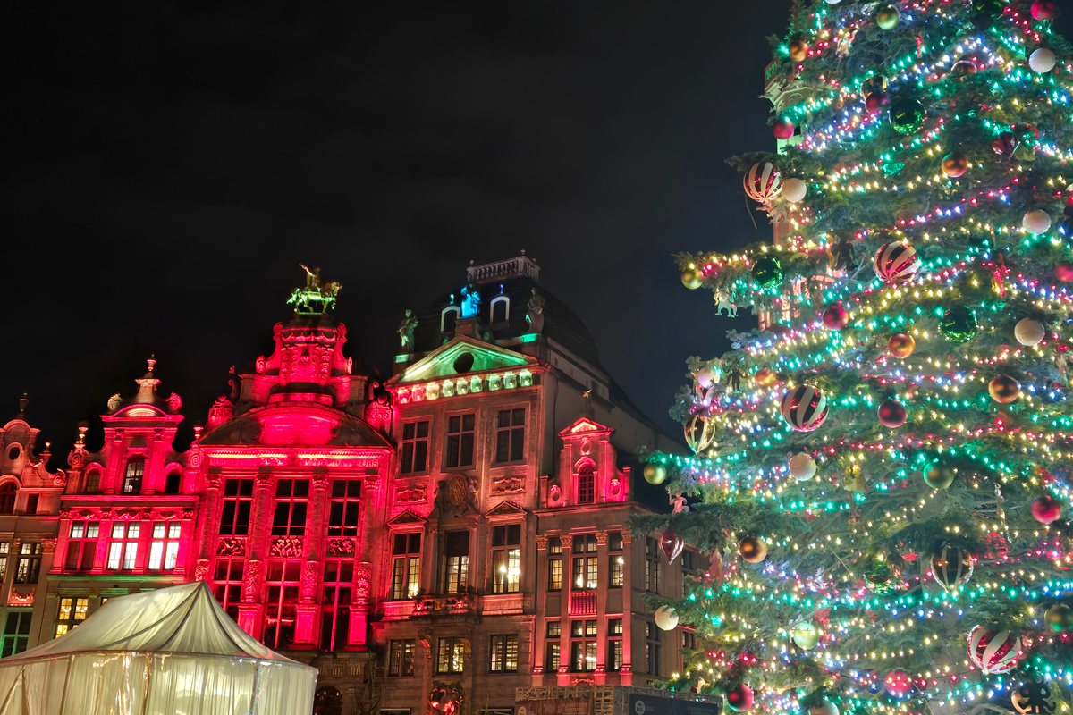 A tall Christmas tree hung with multicoloured lights and striped baubles stands beside ornate guild house facades lit in red and pink on the Grand-Place in Brussels at night, with a white tent in the lower left corner.