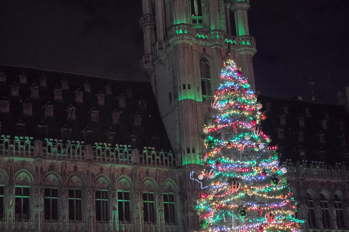 Brussels Town Hall at night, its Gothic tower and facade lit green, with a tall Christmas tree covered in multicoloured lights and red-and-white striped baubles in front.