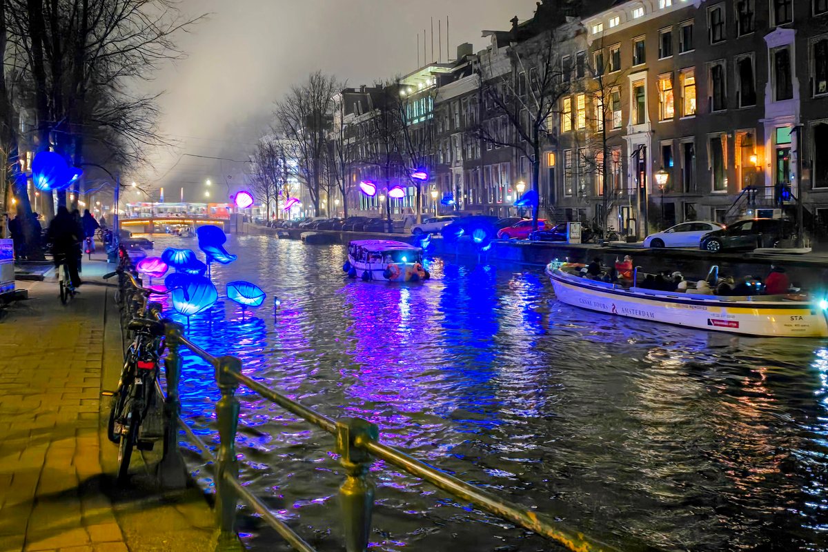 Blue and pink shell-like light sculptures glow above a canal in Amsterdam at night, next to a railing with a parked bicycle, as an open tour boat and a small covered boat pass canal houses with lit windows on a misty evening.