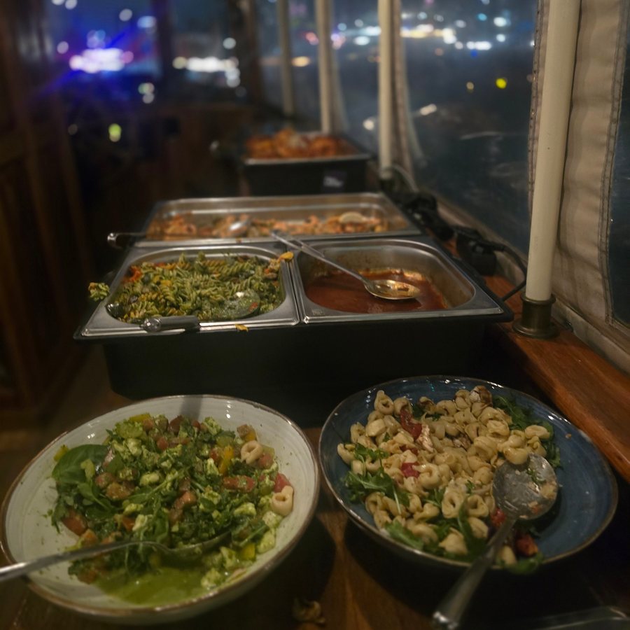 A buffet along the windows of a closed canal boat at night: two salad bowls with rocket in front, one with tortellini, pasta in steel trays behind and city lights outside