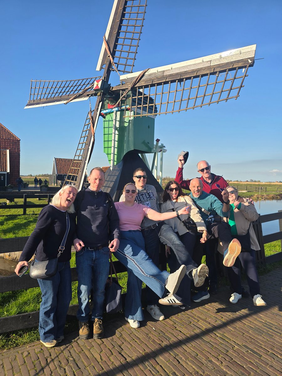 A group of guests laughing and posing in front of a green wooden windmill at Zaanse Schans on a sunny evening, beside a fence and a canal.